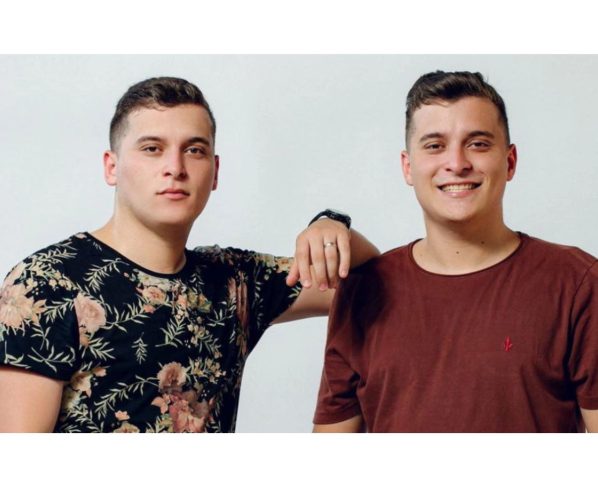 Dois jovens em um fundo claro, um deles usando uma camiseta com estampa floral. Eles sorriem e posam de maneira descontraída, transmitindo uma sensação de amizade e camaradagem. A dupla João e Marcelo agitam o Palco Sunset nesta quinta-feira