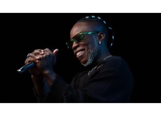 Cantor sorridente segurando um microfone, usando óculos escuros e com estilo moderno em performance ao vivo. Artista Congolês Wugala Flama sobe ao Palco Sunset nesta sexta-feira