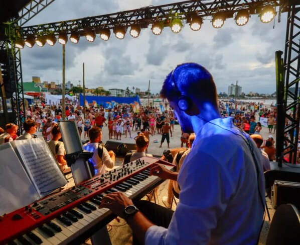Músico tocando teclado em um palco durante um show na praia, com uma grande multidão se divertindo ao fundo sob um céu nublado. Confira as atrações do Palco Sunset para este fim de semana no litoral do Paraná
