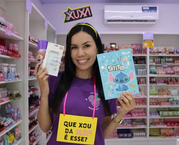 Mulher sorridente segurando produtos de beleza e um caderno com Stitch em uma loja de cosméticos, com destaque para o tema da Xuxa. Confira dicas para brilhar com abadás e acessórios no Carnaval