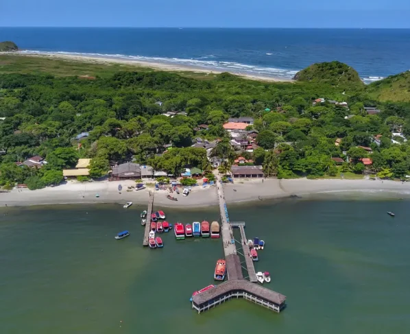 Vista aérea de uma praia tranquila com um píer de madeira, cercado por barcos coloridos e vegetação abundante. Cielo azul e mar calmo. Novo marco regulatório promove preservação e futuro sustentável para a Ilha do Mel