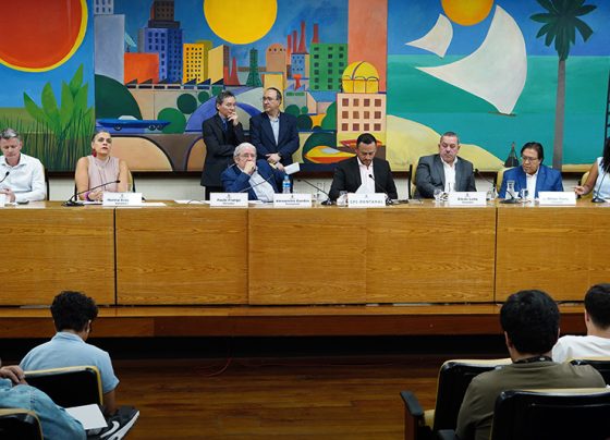 Esta imagem é uma fotografia que captura a reunião de instalação no dia 4 de setembro de 2025 da CPI que investigará as enchentes do Jardim Pantanal e região. Na mesa principal, há os sete vereadores que compõem o colegiado, todos usando trajes formais, que incluem camisas, paletós e, em alguns casos, gravatas. Os nomes dos parlamentares estão indicados em placas à sua frente, mas seus nomes não podem ser identificados por mim. A expressão dos vereadores varia de pensativa a concentrada, e algumas estão interagindo entre si. Atrás da mesa, há duas pessoas de pé conversando. A composição é equilibrada, com atenção centralizada nos indivíduos sentados. O ambiente é o Auditório Prestes Maia, que possui um grande mural colorido ao fundo, o que adiciona vivacidade ao espaço. O mural apresenta um cenário urbano colorido com elementos naturais, como palmeiras e o sol. A iluminação é clara e uniforme, destacando detalhes das expressões faciais dos presentes e dos elementos do mural. As pessoas na reunião aparecem parcialmente na parte inferior da imagem, com atenção voltada para a mesa principal. A imagem, de alta qualidade e bem iluminada, transmite um ambiente profissional e uma atmosfera de discussão séria.