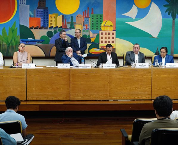 Esta imagem é uma fotografia que captura a reunião de instalação no dia 4 de setembro de 2025 da CPI que investigará as enchentes do Jardim Pantanal e região. Na mesa principal, há os sete vereadores que compõem o colegiado, todos usando trajes formais, que incluem camisas, paletós e, em alguns casos, gravatas. Os nomes dos parlamentares estão indicados em placas à sua frente, mas seus nomes não podem ser identificados por mim. A expressão dos vereadores varia de pensativa a concentrada, e algumas estão interagindo entre si. Atrás da mesa, há duas pessoas de pé conversando. A composição é equilibrada, com atenção centralizada nos indivíduos sentados. O ambiente é o Auditório Prestes Maia, que possui um grande mural colorido ao fundo, o que adiciona vivacidade ao espaço. O mural apresenta um cenário urbano colorido com elementos naturais, como palmeiras e o sol. A iluminação é clara e uniforme, destacando detalhes das expressões faciais dos presentes e dos elementos do mural. As pessoas na reunião aparecem parcialmente na parte inferior da imagem, com atenção voltada para a mesa principal. A imagem, de alta qualidade e bem iluminada, transmite um ambiente profissional e uma atmosfera de discussão séria.