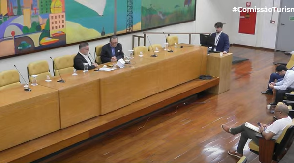 A imagem é uma fotografia de uma reunião da Comissão de Turismo da Câmara Municipal de São Paulo, realizada no dia 9 de setembro de 2025 no Auditório Prestes Maia. Com expressões focadas, dois homens estão sentados atrás de uma longa mesa de madeira, com microfones à disposição. Um deles – de terno escuro e camisa clara - é o vereador Gilberto Nascimento, presidente do colegiado.  No canto direito, um homem em pé, também de terno, está próximo a um púlpito. A hashtag #ComissãoTurismo aparece no canto superior direito. O ambiente é amplo e bem iluminado, com paredes decoradas por um grande mural colorido que ilustra uma paisagem abstrata. O piso é de madeira brilhante, e as cadeiras são estofadas em bege. No fundo, algumas pessoas estão sentadas, aparentando atenção ao que é discutido. A qualidade da imagem é nítida e captura detalhes do cenário e dos participantes.
