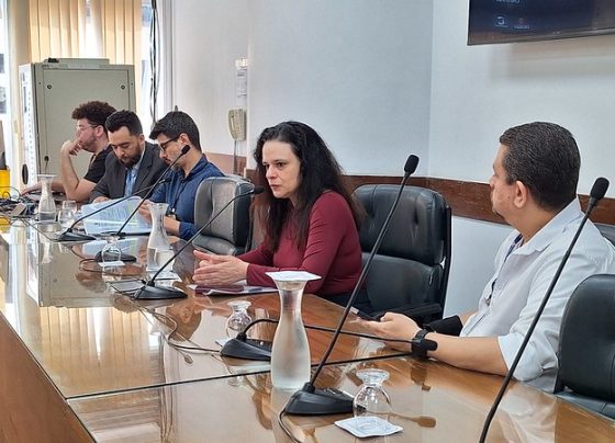 Esta imagem é uma fotografia que captura uma mesa de conferência onde cinco pessoas estão posicionadas lado a lado. Eles estão sentados em cadeiras com microfones à frente, sugerindo que participam de uma discussão ou apresentação. A mulher ao centro, com cabelos longos e escuros, veste uma blusa vermelha e parece estar falando, com uma expressão atenta e gesticulando levemente com as mãos. Os homens ao redor também parecem concentrados, com dispositivos como laptops e documentos à frente. As cores predominantes na imagem são tons neutros, com destaque para o marrom da mesa e o preto das cadeiras. O ambiente é uma sala de conferências clara, com paredes brancas e uma grande mesa de madeira que se estende horizontalmente. No fundo, cortinas verticais suavemente fechadas permitem a entrada de luz natural, contribuindo para a iluminação suave da cena. Vista lateralmente, a imagem mostra ainda pequenos objetos como garrafas de vidro e copos sobre a mesa. A fotografia é bem iluminada, com alta qualidade, capturando os detalhes de forma nítida, como as expressões faciais dos participantes e os materiais utilizados. Essa descrição sugere que o contexto da imagem é uma reunião ou seminário profissional, transmitindo um ambiente de concentração e colaboração.