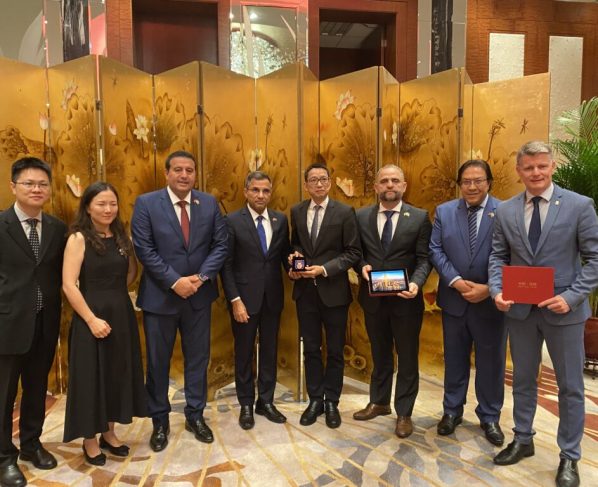 Foto de grupo com oito homens de terno e uma mulher de vestido preto, todos em pé lado a lado, alguns seguram objetos. Fundo com biombo dourado de flores, iluminação interna quente, ambiente refinado e solene.