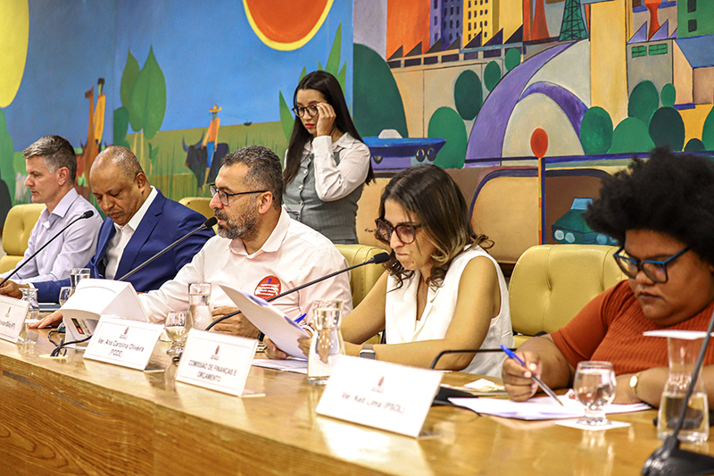 Foto mostra cinco vereadores da Comissão de Finanças e Orçamento, em reunião realizada no dia 10 de setembro de 2025 na Câmara Municipal de São Paulo. Todos estão sentados em uma mesa de reunião do Auditório Prestes Maia, lendo documentos e escrevendo. Uma mulher em pé atrás atende celular. Ao fundo, mural colorido com formas geométricas e paisagem urbana. Sala iluminada, mesa de madeira com microfones e copos de água.