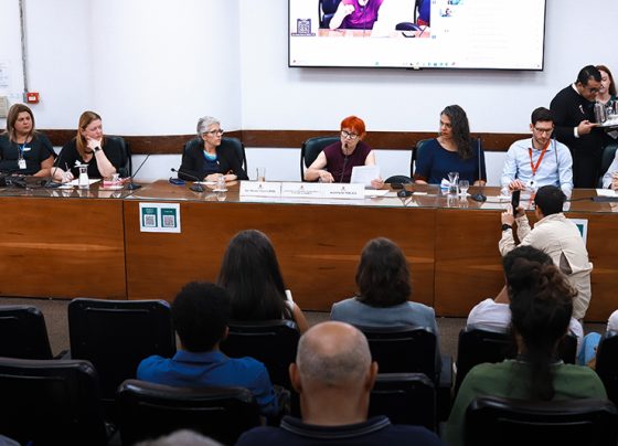 Imagem mostra uma mesa com sete pessoas sentadas em frente a um público. Uma mulher ao centro fala ao microfone. Sala de reunião iluminada, parede branca e tela projetada ao fundo. Ambiente formal e organizado.