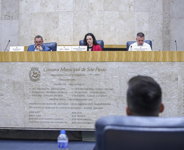 Foto da reunião da CPI da Íris, de 30 de setembro de 2025 na Câmara Municipal de São Paulo; três vereadores sentados à mesa central, microfones e placas de identificação. Fundo de mármore claro com nomes gravados; ambiente iluminado, tom formal e institucional. No primeiro plano, depoente sentado em uma das cadeiras do Plenário 1º de Maio.