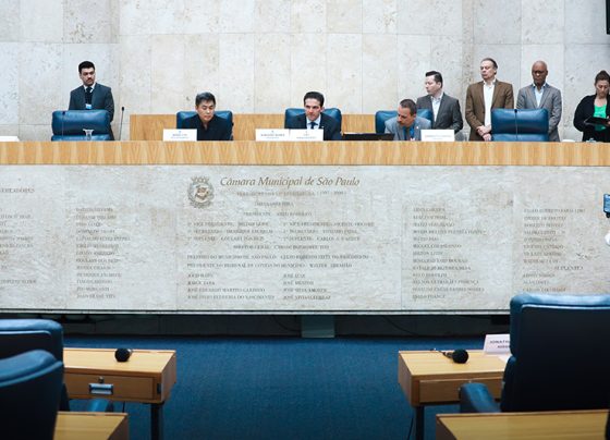 Reunião da CPI dos Pancadões, realizada em 30 de outubro de 2025. Imagem do Plenário da Câmara Municipal de São Paulo com três vereadores sentados atrás de uma bancada de mármore clara. Em pé, ao fundo, servidores da Casa. A parede é bege, lisa e tem e a bancada de mármore tem o brasão da Câmara. Iluminação branca e ambiente formal de reunião.