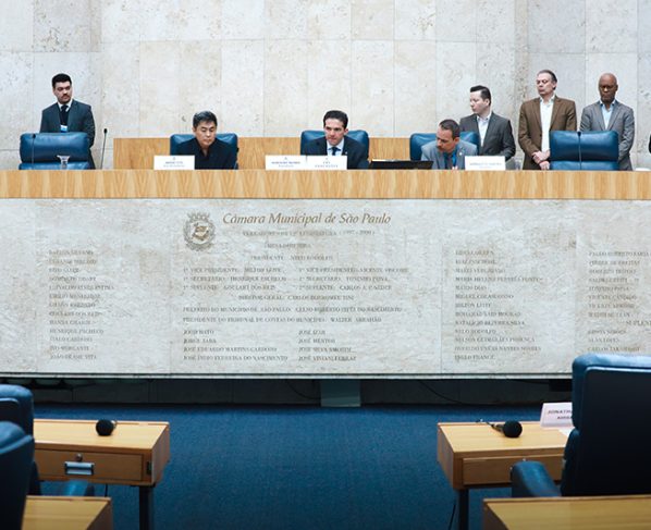 Reunião da CPI dos Pancadões, realizada em 30 de outubro de 2025. Imagem do Plenário da Câmara Municipal de São Paulo com três vereadores sentados atrás de uma bancada de mármore clara. Em pé, ao fundo, servidores da Casa. A parede é bege, lisa e tem e a bancada de mármore tem o brasão da Câmara. Iluminação branca e ambiente formal de reunião.