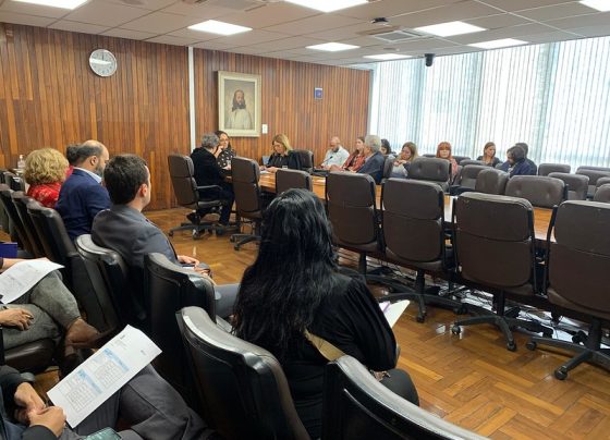 Reunião em sala com mesa longa e pessoas sentadas discutindo documentos. Parede de madeira ao fundo com quadro e relógio. Ambiente interno iluminado por luz natural das janelas com persianas. Clima formal de reunião.