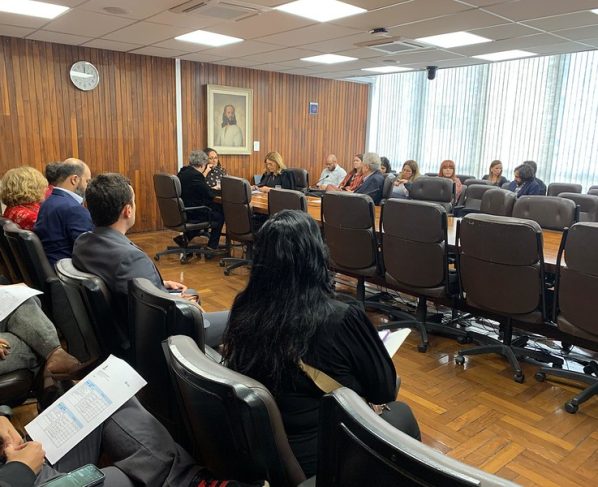 Reunião em sala com mesa longa e pessoas sentadas discutindo documentos. Parede de madeira ao fundo com quadro e relógio. Ambiente interno iluminado por luz natural das janelas com persianas. Clima formal de reunião.