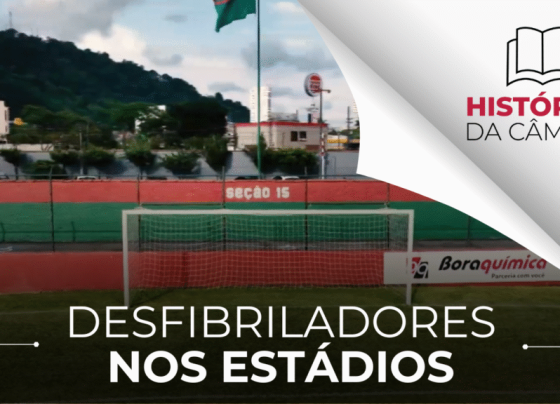 A imagem é uma fotografia de um estádio de futebol. Há uma trave central branca com uma rede e uma parede ao fundo com as cores vermelha e verde, indicando "seção 15". Ao fundo, há vegetação e prédios, com uma bandeira no alto. No canto direito, há o logotipo "HISTÓRIAS DA CÂMARA" e as palavras "DESFIBRILADORES NOS ESTÁDIOS". A iluminação é natural e clara.