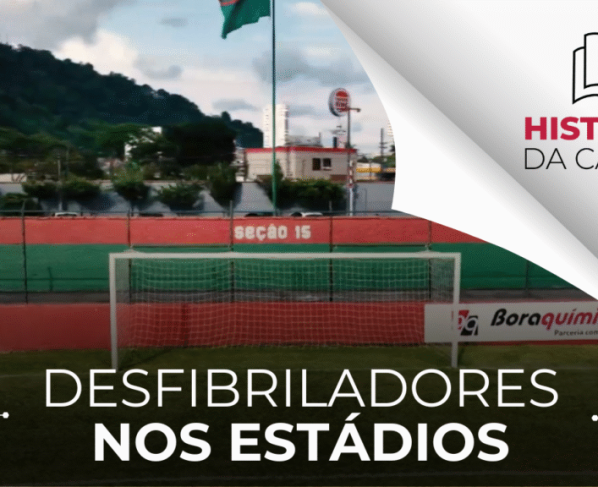A imagem é uma fotografia de um estádio de futebol. Há uma trave central branca com uma rede e uma parede ao fundo com as cores vermelha e verde, indicando "seção 15". Ao fundo, há vegetação e prédios, com uma bandeira no alto. No canto direito, há o logotipo "HISTÓRIAS DA CÂMARA" e as palavras "DESFIBRILADORES NOS ESTÁDIOS". A iluminação é natural e clara.
