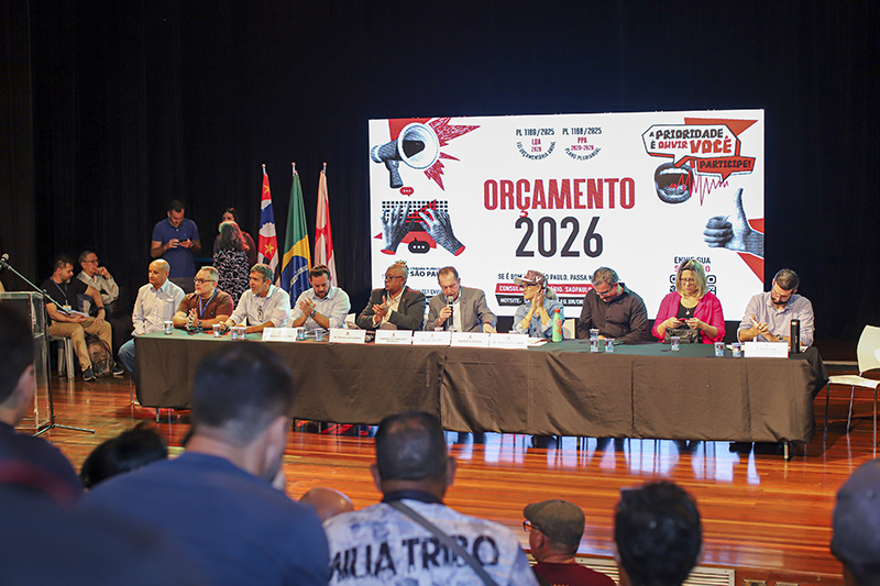 Imagem da Audiência Pública regional sobre o orçamento 2026 e o PPA, realizada no CEU Paz, zona norte, em 18 de outubro de 2025. A foto do evento mostra com nove pessoas sentadas à mesa - entre vereadores e outras autoridades - sobre palco, diante de público. Atrás, painel grande com texto. Banner com fundo branco e vermelho traz o texto “Orçamento 2026” e “A prioridade é ouvir você. Participe!”. Ambiente interno iluminado.