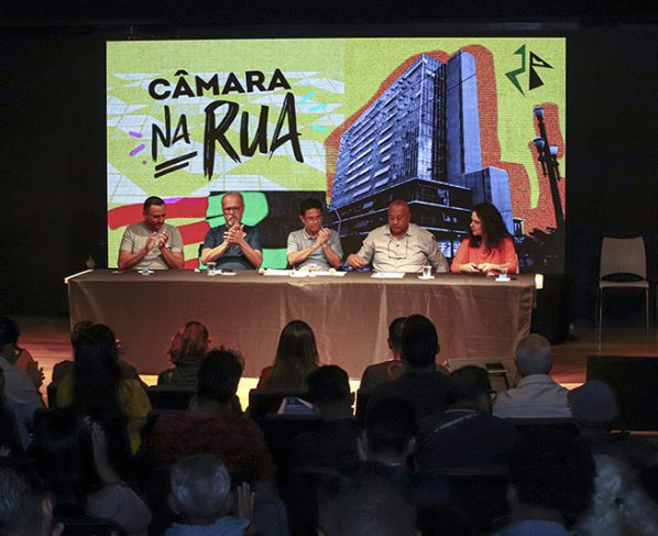 Câmara na Rua, realizado no CEU Paraisópolis em 1 de novembro de 2025. Cinco vereadores sentados à mesa em auditório, diante de público. Atrás, tela mostra “Câmara na Rua” e imagem de prédio. Ambiente interno iluminado, fundo colorido e palco escuro. Cena de evento público oficial.