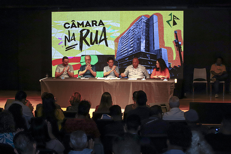 Câmara na Rua, realizado no CEU Paraisópolis em 1 de novembro de 2025. Cinco vereadores sentados à mesa em auditório, diante de público. Atrás, tela mostra “Câmara na Rua” e imagem de prédio. Ambiente interno iluminado, fundo colorido e palco escuro. Cena de evento público oficial.