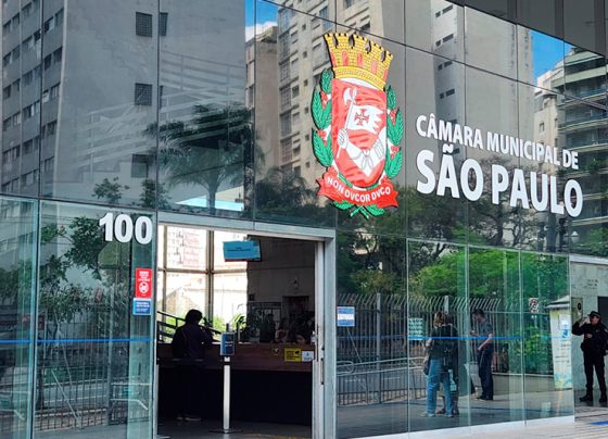 Fachada de vidro com brasão e texto “Câmara Municipal de São Paulo” e número 100. Pessoas próximas à entrada. Prédio urbano espelhado, refletindo árvores e construções. Luz natural, dia claro, imagem nítida.