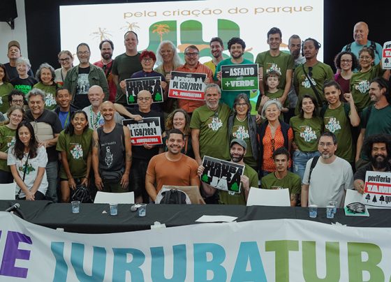 Audiência da Comissão de Administração Pública em Escola no bairro do Socorro, zona sul, sobre a criação do Parque Jurubatuba. Foto de grupo com cerca de 30 pessoas sorrindo, muitas vestem camisetas verdes com o logo “JURUBATUBA”. Algumas seguram cartazes. Ambiente interno iluminado, painel ao fundo com texto “Pela criação do parque JURUBATUBA”. Mesa à frente com faixa azul e verde.
