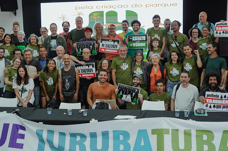 Audiência da Comissão de Administração Pública em Escola no bairro do Socorro, zona sul, sobre a criação do Parque Jurubatuba. Foto de grupo com cerca de 30 pessoas sorrindo, muitas vestem camisetas verdes com o logo “JURUBATUBA”. Algumas seguram cartazes. Ambiente interno iluminado, painel ao fundo com texto “Pela criação do parque JURUBATUBA”. Mesa à frente com faixa azul e verde.