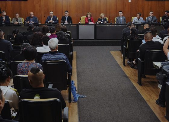 Imagem mostra uma mesa com nove pessoas sentadas em sessão formal, diante de plateia em auditório. Ambiente com painéis de madeira, iluminação interna uniforme e clima de reunião oficial.