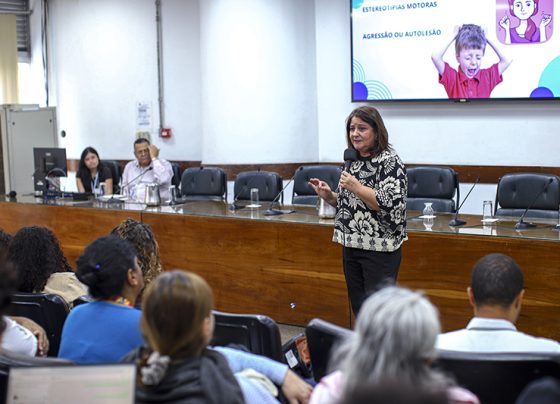 Mulher em pé fala ao público segurando microfone em sala com mesa e telão. Pessoas sentadas observam. Fundo com parede branca e projeção escrita “Estereotipias motoras. Agressão ou autolesão”. Iluminação interna clara.