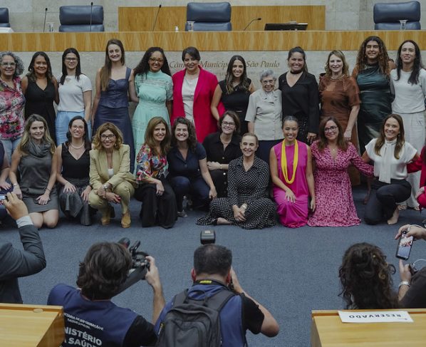 Foto colorida de grupo com 22 mulheres, em duas fileiras, posando no plenário da Câmara Municipal de São Paulo. Ao fundo, bancada de madeira clara e cadeiras azuis; iluminação interna uniforme e ambiente formal.
