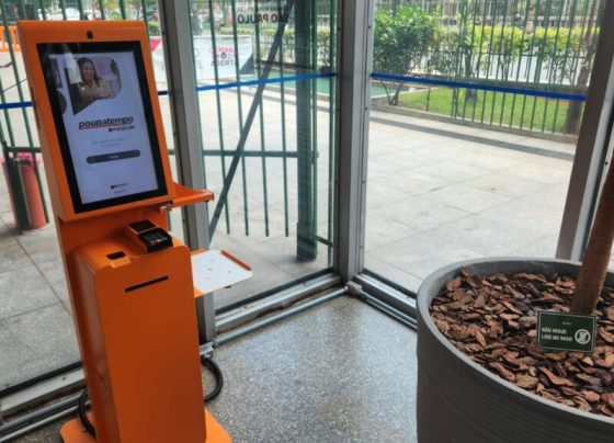Totem de autoatendimento laranja do Poupatempo com tela e leitor de cartão. Ao lado, grande vaso com plaqueta “NÃO JOGUE LIXO NO VASO”. Ambiente interno com piso de granito claro e janelas de vidro que mostram área externa iluminada e cercada.