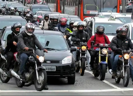 Motociclistas e carros em trânsito intenso em avenida urbana, com ônibus e táxis ao fundo, ilustrando frota de veículos em Alagoas