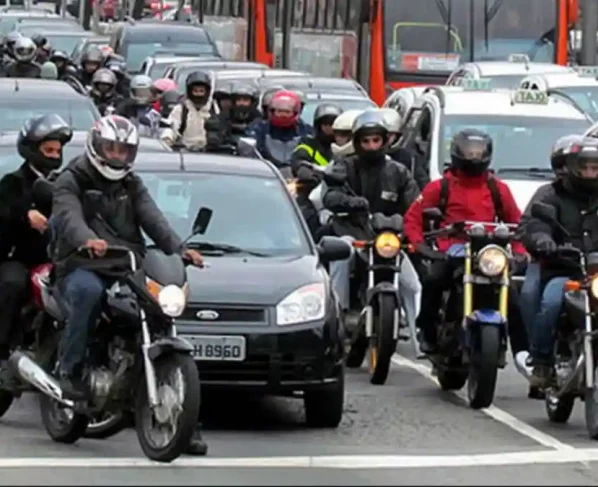 Motociclistas e carros em trânsito intenso em avenida urbana, com ônibus e táxis ao fundo, ilustrando frota de veículos em Alagoas
