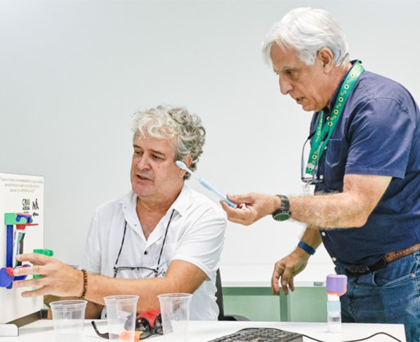Duas pessoas em sala de aula manipulam um modelo científico colorido sobre a mesa, com copos plásticos e ferramentas ao redor.