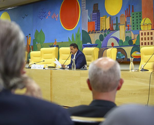 Imagem de reunião em plenário com homem falando ao microfone em mesa de madeira clara e cadeiras amarelas. Ao fundo, painel colorido com formas geométricas e paisagem urbana sob luz interna clara.