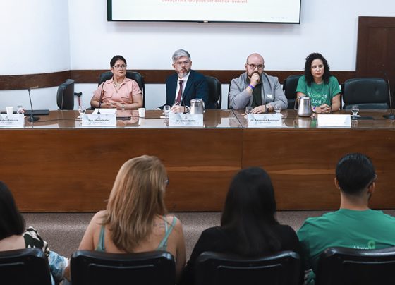 Imagem de uma mesa de audiência com quatro pessoas sentadas, três homens e uma mulher usam trajes formais. Ao fundo, parede branca e painel de projeção; público sentado observa, iluminação clara e ambiente institucional.