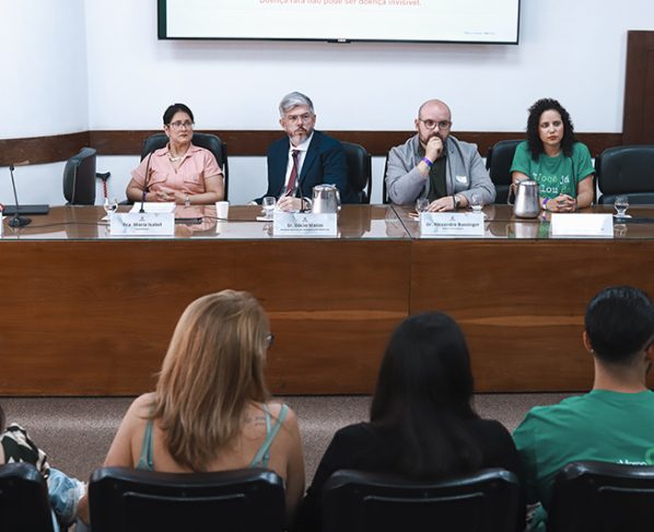Imagem de uma mesa de audiência com quatro pessoas sentadas, três homens e uma mulher usam trajes formais. Ao fundo, parede branca e painel de projeção; público sentado observa, iluminação clara e ambiente institucional.