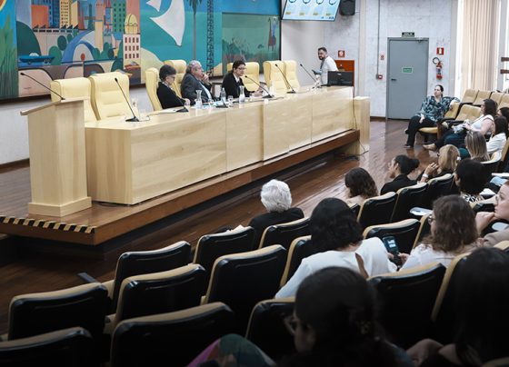 Foto de auditório com quatro pessoas sentadas à mesa em frente a microfones e público assistindo. Fundo com painel colorido e iluminação natural; ambiente formal e bem iluminado.