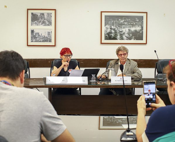 Imagem de reunião com duas pessoas sentadas à mesa, microfones e placas de identificação à frente. Sala de conferência iluminada; fundo com fotos emolduradas em preto e branco nas paredes.