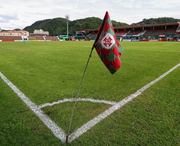 Bandeirinha de escanteio com escudo da Portuguesa Santista em campo de futebol, com arquibancadas ao fundo.