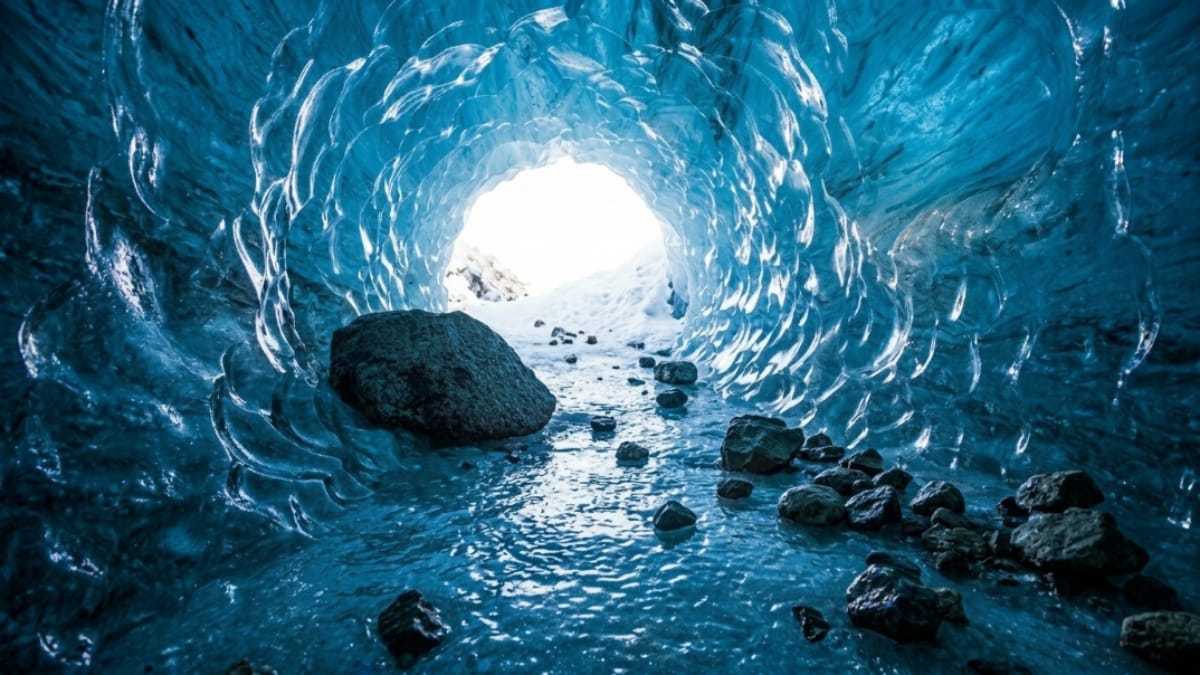 Vista do interior de uma caverna de gelo azul com paredes de textura ondulada e translúcida. O chão está coberto por gelo e pequenas pedras escuras, levando a uma abertura luminosa ao fundo que revela uma paisagem externa com neve.