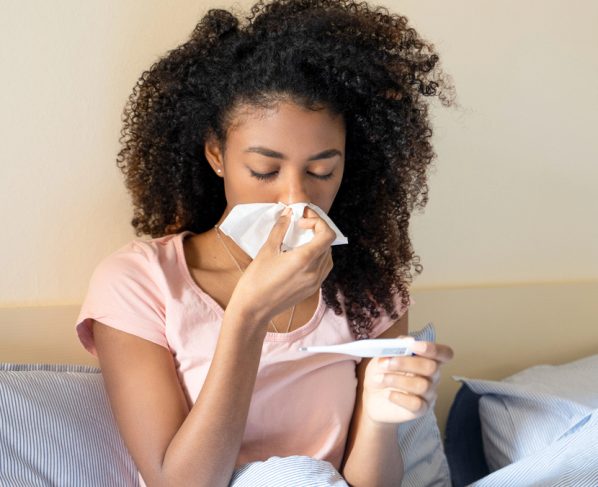 mulher de cama, com gripe, assoando o nariz e olhando o termômetro