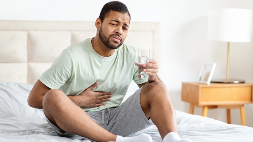 homem sentado na cama, com copo d'água numa mão e outra no abdômen, em sinal de dor