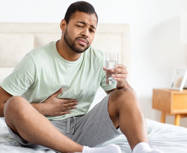 homem sentado na cama, com copo d'água numa mão e outra no abdômen, em sinal de dor