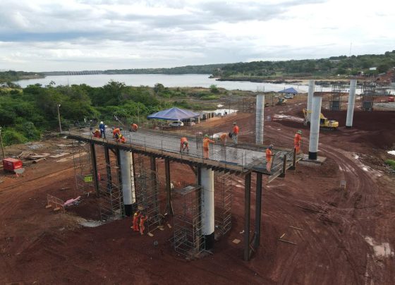 Construction workers in orange safety gear assemble an elevated steel walkway on concrete columns above a muddy site beside a river with green hills in the background and tents nearby.