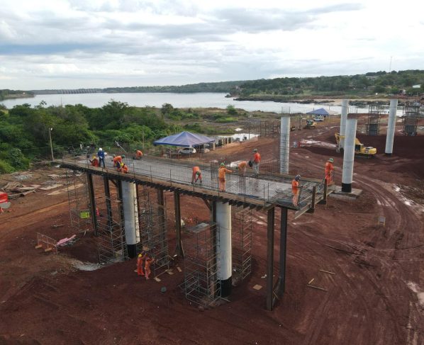 Construction workers in orange safety gear assemble an elevated steel walkway on concrete columns above a muddy site beside a river with green hills in the background and tents nearby.