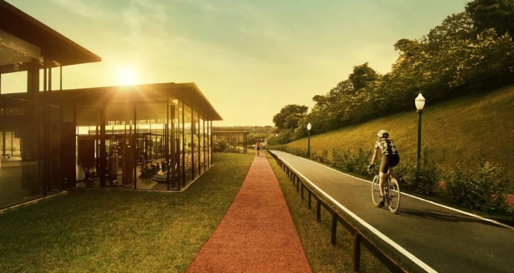 A imagem mostra uma área de lazer ao ar livre com clima de fim de tarde. Há uma ciclovia ao lado de uma pista de caminhada, com uma pessoa andando de bicicleta. À esquerda, aparece um espaço moderno com paredes de vidro, provavelmente uma academia. O cenário é cercado por natureza, com gramados e árvores, transmitindo tranquilidade e qualidade de vida.
