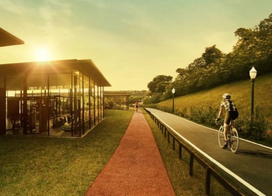 A imagem mostra uma área de lazer ao ar livre com clima de fim de tarde. Há uma ciclovia ao lado de uma pista de caminhada, com uma pessoa andando de bicicleta. À esquerda, aparece um espaço moderno com paredes de vidro, provavelmente uma academia. O cenário é cercado por natureza, com gramados e árvores, transmitindo tranquilidade e qualidade de vida.