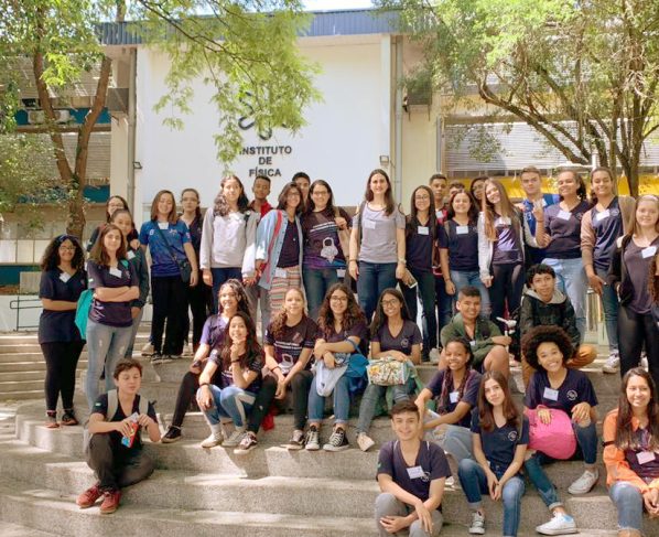 Grupo de estudantes, alguns em pé e outros sentados nos degraus de uma escadaria, em frente a um prédio baixo