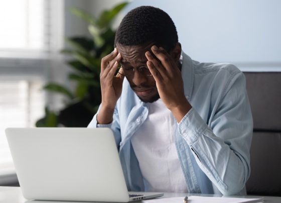 homem jovem de frente ao computador, em escritório, com a mão na cabeça tentando se lembrar de algo