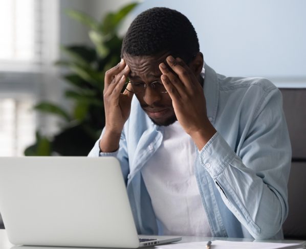 homem jovem de frente ao computador, em escritório, com a mão na cabeça tentando se lembrar de algo