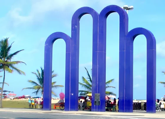 Monumento dos Arcos da Orla de Atalaia, em Aracaju, com coqueiros e praia ao fundo.